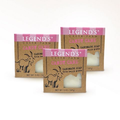 Three packages of Legend's Creek Farm Candy Cane Handmade Goat Milk Soap, with the scent of candy canes. The packaging is primarily pink and beige, with an illustration of a goat and candy cane motifs.